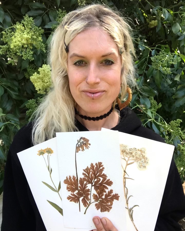 A blonde woman is smiling, looking at the camera while holding up a piece of paper with flowers she pressed showing.